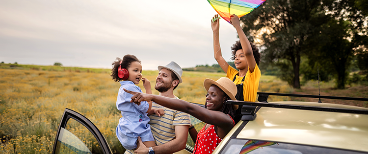 A happy family looks into the trunk of their car. The father hands the mother an orange while the child reaches for something in the trunk.